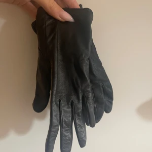 Svarta skinnhandskar från H&M - Svarta handskar i mjukt skinn från H&M, klassisk och stilren design med slät yta och diskreta sömmar. Handskarna har fem fingrar och är perfekta för kalla dagar. Storlek S enligt etiketten. Knappt använda, toppskick.