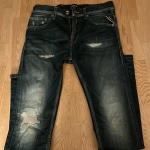 Replay jeans med slitningar, midja 28/30 - Snygga mörkblå Replay jeans med slitna detaljer och lagade hål på benen. Klassisk femficksmodell med dragkedja och unika bakfickor med kontrastsömmar. Jeansen har en cool tvätt och är tillverkade i bomull för en skön känsla. Sitter som Super slim/skinny