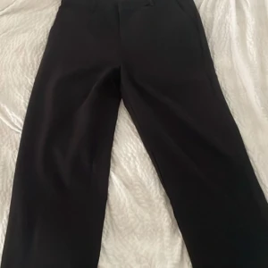 Svarta kostymbyxor med raka ben - Snygga svarta kostymbyxor med raka ben och klassiska hällor i midjan. Byxorna har diskreta fickor bak och är perfekta för en clean och stilren look. Materialet känns slätt och dressat, passar dig som gillar en enkel men uppklädd vibe. 