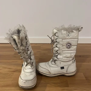 Vita vinterstövlar med fuskpäls - Säljer ett par vita vinterstövlar från leat med snörning och fluffig fuskpäls upptill. Stövlarna har rund tå, platt sula och är fodrade för extra värme. Perfekta för kalla dagar och har en sportig look med detaljerade sömmar. Är lite slitna framtill men är absolut inget man tänker på därför skulle jag säga att det är i mycket bra skick!!