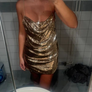 Glittrig guldklänning från ASOS - Säljer en glittrig kortklänning från ASOS i guld med paljetter över hela klänningen. Klänningen har tunna axelband, snygg draperad front och snörning i ryggen för extra detalj. Perfekt för dig som vill sticka ut och glänsa.