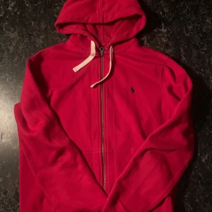  Ralph Lauren Zip hoodie  - Riktigt snygg röd hoodie från Polo Ralph Lauren med dragkedja och vit snörning i huvan. Klassisk broderad logga på bröstet. Tillverkad i mjuk bomullsmix med polyester för extra komfort. Perfekt för chill dagar eller när du vill ha en stilren streetlook.