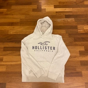Vit hoodie från Hollister M - Vit hoodie från Hollister med stor logga och text på bröstet. Tröjan har huva med snörning och är tillverkad i mjuk bomullsblandning. Perfekt för chill dagar och snygg till jeans eller joggers.
