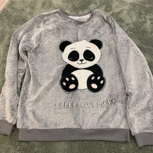 Grå fluffig tröja med panda från Cubus - Supermjuk och fluffig grå tröja från Cubus med en söt panda framtill och texten 'SLEEP ALL DAY'. Tröjan har långa ärmar, ribbade muddar och är perfekt för mysiga dagar hemma. Materialet är fleece och motivet är broderat för extra detalj.