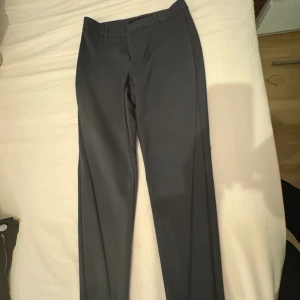 Grå kostymbyxor från Vero Moda - Snygga grå kostymbyxor från Vero Moda med klassisk rak passform och diskreta fickor fram. Byxorna har bälteshällor och är perfekta för en clean och stilren look. Materialet känns mjukt och följsamt, vilket gör dem bekväma hela dagen.