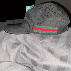 Svart Gucci keps - Svart keps från Gucci med klassiskt GG-monogram över hela ytan och en grön-röd rand på sidan. Kepsen har böjd skärm och justerbar rem baktill. Materialet är bomull och polyester, vilket gör den både snygg och bekväm. Perfekt för dig som vill ha en lyxig streetstyle.