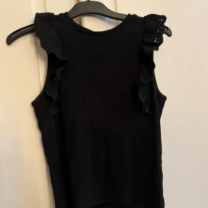 Svart ärmlös topp med volang från Kappahl - Svart ribbad topp från Kappahl i storlek 158/164. Ärmlös modell med breda axelpartier dekorerade med broderade volanger som har små hålmönster. Perfekt för dig som gillar stilrena detaljer och vill ha något som sticker ut lite extra.