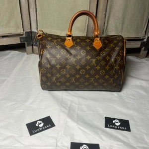 Louis Vuitton Speedy 30 - Klassisk handväska från Louis Vuitton i brunt skinn med ikoniskt monogrammönster i gult. Väskan har rund form, två robusta handtag och dragkedja upptill. Perfekt för dig som vill ha en tidlös och lyxig accessoar med plats för det viktigaste.