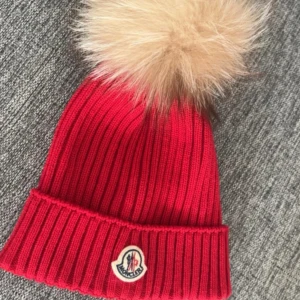 Röd Moncler mössa med pälsboll - Ribbstickad röd mössa från Moncler med stor beige pälsboll på toppen och klassisk Moncler-logga framtill. Perfekt accessoar för kalla dagar. Liten och tight i storleken!!