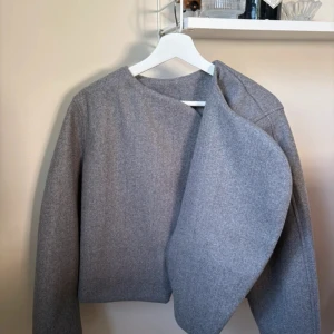 Grå omlott kappa med oversized passform - Säljer en stilren grå kappa med omlott-design och bred krage. Jackan har en loose och oversized passform som ger en cool och avslappnad vibe. Materialet är mjukt och känns som ull, perfekt för höst och vinter. Långärmad och enkel att matcha med olika outfits.