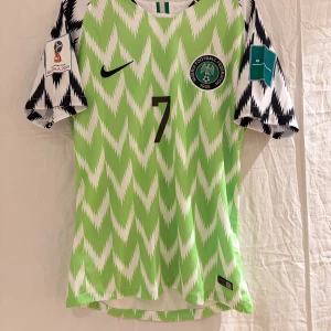 Nigeria VM 2018 fotbollströja Musa #7 - Säljer en retro Nigeria VM 2018 fotbollströja från Nike med Musa och nummer 7 på ryggen. Tröjan är grön och vit med ett unikt zigzag-mönster, korta ärmar och autentiska VM-patchar. Materialet är lätt och ventilerande polyester, perfekt för match eller samling.