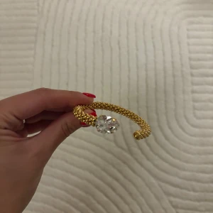 Guldarmband Caroline Svedbom med kristall - Unikt guldarmband från Caroline Svedbom med en stor, droppformad klar kristall. Armbandet har en cool pärlstruktur och är öppet i modellen, vilket gör det enkelt att ta på. Perfekt statement piece för dig som gillar bling och exklusiv design.