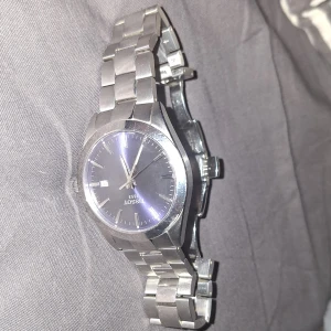 Tissot klocka med blå urtavla - Stilren Tissot armbandsklocka i silverfärgat stål med länkarmband och blå urtavla. Klockan har datumvisning vid klockan tre och tydliga indexmarkeringar. Klassisk rund boett och robust känsla, perfekt för dig som gillar tidlös design.