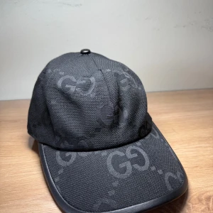 Svart keps från Gucci med GG-mönster - Snygg svart keps från Gucci med ikoniskt GG-mönster ton-i-ton över hela kepsen. Kepsen har böjd skärm med läderdetalj runt kanten och justerbar rem baktill. Tillverkad i slitstark bomull och polyester, perfekt för dig som vill ha en lyxig streetstyle-look.