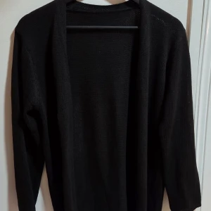 Svart stickad kofta med lång ärm, M - En svart, enkel och stilren stickad kofta med lång ärm. Koftan är öppen framtill utan knappar och har en rak, lite loose passform. Perfekt att slänga över en t-shirt eller topp för en chill look. Ca 78 cm lång.