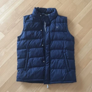 Mörkblå dunväst från GANT - Snygg mörkblå dunväst från GANT. Västen har dragkedja och tryckknappar framtill, samt två fickor på sidorna. Diskret GANT-logga broderad på bröstet. Perfekt för lager-på-lager under kyliga dagar. Storleken är för 176 cm och motsvarar storlek S