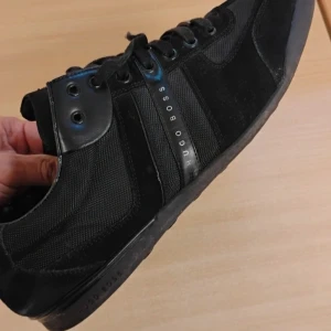 Svarta sneakers från Hugo Boss - Snygga svarta sneakers från Hugo Boss med stilren design. Skorna har snörning, rund tå och detaljer i både mocka och skinn. Sidorna pryds av Hugo Boss-loggan och hälen har ribbad skinnpanel. Perfekta för dig som gillar klassisk streetstil.