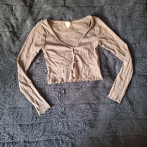 Beige croppad ribbad blus med knappar - Säljer en beige croppad blus med lång ärm och v-ringning. Toppen har ribbad struktur, små knappar framtill och vågig kant vid nederdel och ärmslut. Perfekt för dig som gillar stilrena och enkla plagg med en twist.