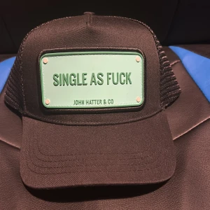 Svart keps John Hatter & Co - Svart keps från John Hatter & Co med mesh på sidorna och justerbar snapback. Framtill sitter en mintgrön skylt med texten 'SINGLE AS FUCK'. Kepsen har broderad logga på baksidan och insidan pryds av texten 'MAKE A STATEMENT'. Perfekt för dig som vill sticka ut.