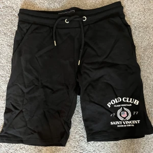 Svarta shorts från Polo Club Saint Vincent - Svarta shorts från Polo Club Saint Vincent med tryck framtill och snörning i midjan. De har en normal passform, sidofickor och en bakficka. Materialet är mjuk bomull, perfekt för chill dagar. Klassisk look med ribbad midja och stilren design.