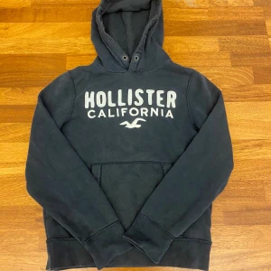 Svart hoodie från Hollister  - Svart hoodie från Hollister med stor vit logga och texten 'Hollister California' på bröstet. Klassisk känguruficka framtill och huva med dragsko. Mjuk insida och långa ärmar, perfekt för kyliga dagar. Den passar som en M