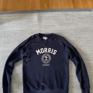 Mörkblå sweatshirt från Morris - Säljer en mörkblå sweatshirt från Morris med vit logga och tryck framtill. Tröjan har rund halsringning, långa ärmar och ribbade muddar vid ärmslut och nederkant. Perfekt för en avslappnad och sportig stil.