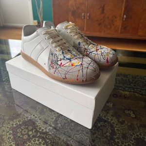 Maison Margiela  färgstänk - Maison Margiela sneakers i vitt skinn med beige mockadetaljer och coolt färgstänksmönster i blått, rött, gult och svart. Klassisk rund tå, platt gummisula och snörning. Snygg design som sticker ut och passar dig som gillar unika sneakers.