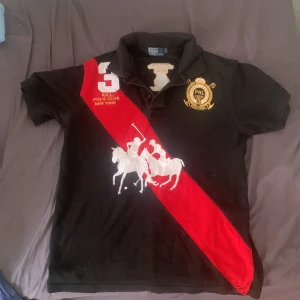 Svart pikétröja Polo Ralph Lauren - Svart pikétröja från Polo Ralph Lauren med röd diagonal rand, stora broderade polospelare i vitt och guldiga detaljer. Klassisk krage, kort ärm och broderad logga på bröstet. Snygg och sportig stil med tydlig New York-inspiration.