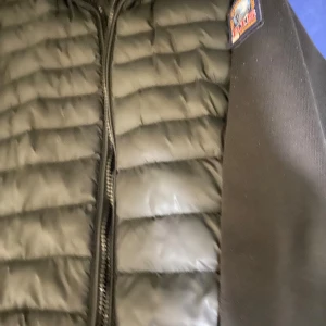 Svart pufferjacka från Parajumpers - Svart pufferjacka från Parajumpers med quiltad front och stickade ärmar. Jackan har dragkedja med orange detalj, ribbade muddar och klassisk Parajumpers-patch på axeln. Materialet är polyester och bomull, vilket gör den både mjuk och varm.