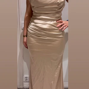 Champagnefärgad långklänning i satin - Elegant axelbandslös långklänning i champagnefärgad satin med draperad överdel och figurnära passform. Klänningen har snygga rynkade detaljer och dragkedja bak för en smickrande siluett. Storlek 40 passar även 36-38! Klänningen är från SE fashion 