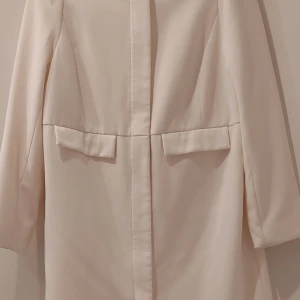 Vit-beige kappa från H&M, stl 44 - Stilren beige kappa från H&M i storlek 44. Lång modell med dold dragkedja framtill och två fickor med lock. Mjuk känsla och klassisk look, perfekt för dig som gillar minimalistisk stil.