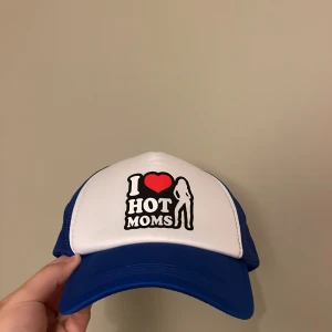 Blå och vit truckerkeps I Love Hot Moms - Snygg truckerkeps med vit front och blå mesh på sidorna och bak. Framsidan har trycket 'I ♥ HOT MOMS' med en siluett. Skärmen är blå och kepsen har klassisk form med justerbar snapback. Perfekt statement piece för dig som vill sticka ut.