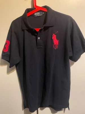 Svart pikétröja från Ralph Lauren - Säljer en svart pikétröja från Ralph Lauren med röd broderad logga på bröstet och en stor röd siffra 3 på ärmen. Tröjan har klassisk krage, två knappar och korta ärmar. Materialet är mjuk bomull och passformen är normal.