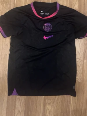 Svart PSG x Nike Dri-Fit t-shirt S - Säljer en svart PSG t-shirt från Nike med Dri-Fit-teknologi. Tröjan har lila och rosa detaljer vid krage och ärmslut, samt ett Paris Saint-Germain-emblem och en rosa Nike-logga med drip-effekt på bröstet. Perfekt för dig som gillar fotboll och streetwear.
