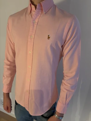 Rosa skjorta från Polo Ralph Lauren - Säljer en slim fit Polo Ralph Lauren skjorta i ljusrosa, i storlek S. Skjortan har klassisk krage med knappar, lång ärm och ikonisk broderad logga på bröstet. 