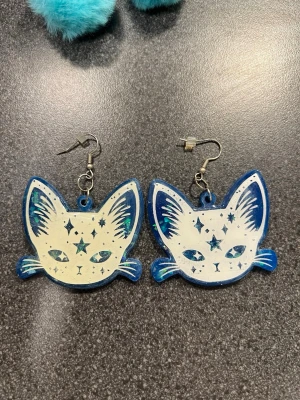 Blå glittriga kattörhängen - Unika örhängen i form av galax kattansikten med blå och vita färger, dekorerade med stjärnor och glitterdetaljer. Framsidan har ett mystiskt kattansikte med stjärnmönster, medan baksidan är helt blå med glittrande inslag. Perfekt för dig som gillar statement-smycken och coola accessoarer. En av kattern har ett mer vitt ansikte och det andra har ett mer varmare ansikte (kolla någrant på bilderna)