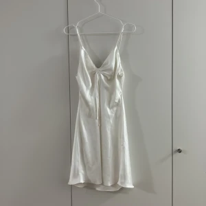 Vit satin klänning från Bik Bok - Säljer en vit kortklänning från Bik Bok i glansigt satinmaterial. Klänningen har smala axelband, v-ringad hals och snygg snörning framtill som ger en cool detalj. Perfekt för dig som gillar stilrena och trendiga plagg. Insydd i midjan så skulle säga att den är M/S