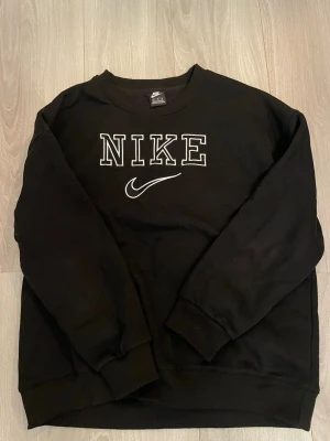 Svart sweatshirt från Nike - Svart sweatshirt från Nike med stort broderat logotyptryck i vitt på bröstet. Tröjan har rund hals, långa ärmar och ribbade muddar vid ärmslut och nederkant. Perfekt för en chill och sportig stil.