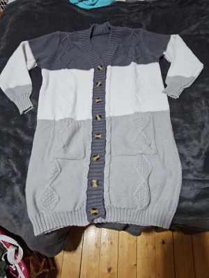 Grå och vit lång kofta med knappar - Säljer en lång kofta i storlek S med breda blockränder i mörkgrått, vitt och ljusgrått. Koftan har stickat mönster, ribbad kant, stora fickor och träknappar framtill. Materialet är 100% polyester och den har långa ärmar. Perfekt för att hålla sig varm och snygg under kyliga dagar.