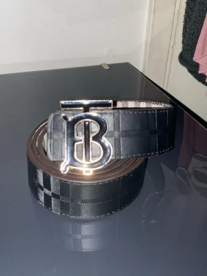 Svart rutig väst från Burberry - Snygg svart väst med diskret rutigt mönster och klassisk Burberry-design. Västen har en stilren form och är tillverkad i ett slitstarkt material som ger en exklusiv känsla. Perfekt för dig som vill ha en modern och trendig look med ikonisk B-detalj framtill.