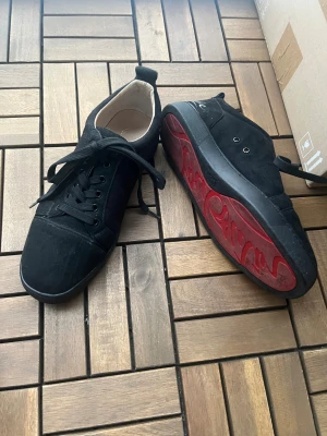 Svarta sneakers från Christian Louboutin - Säljer ett par svarta sneakers från Christian Louboutin med klassisk röd sula och snygg signatur. Skorna har snörning, rund tå och är gjorda i mocka med ljus insida. Perfekt för dig som vill ha en lyxig och stilren look med en ikonisk detalj under foten. 