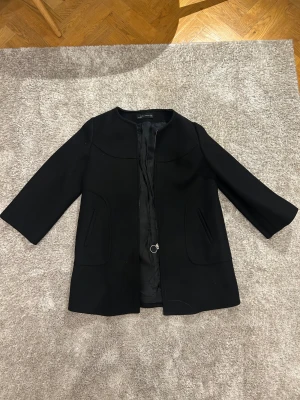 Svart kappa från Zara Woman XL - Svart kappa från Zara Woman i storlek XL. Stilren design med rund hals, raka fickor och en snygg silverfärgad dragkedja med ring. Materialet är ullblandning och kappan har trekvartsärmar. Perfekt för dig som gillar minimalistisk och klassisk stil.