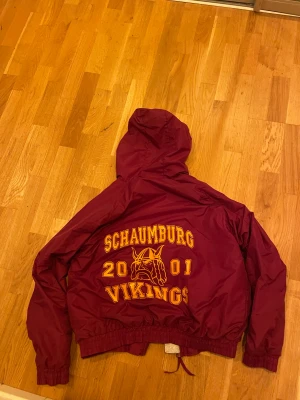 Burgundy vindjacka från Tri-Mountain - Säljer en burgundy vindjacka med huva från Tri-Mountain. Jackan har gul broderad text både fram och bak, med tryck 'Schaumburg Vikings 2001' och ett vikingmotiv på ryggen. Insidan av huvan är ljusgrå och jackan har dragkedja samt resår vid ärmslut och midja.