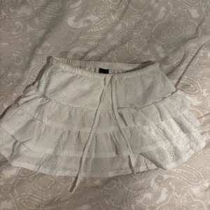 Vit volangkjol med broderi och snörning - Supersöt vit kortkjol med flera volanger och broderade detaljer. Kjolen har snörning som anpassar storleken runt midjan.                                                                                Storlek: S                                                                                    Köpte för 150 kr och säljer för 60 kr.