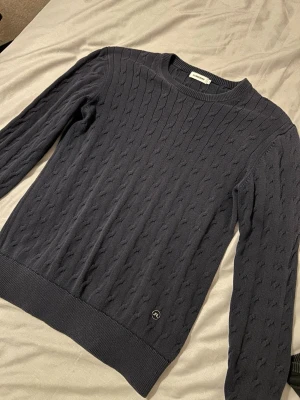 Mörkblå stickad tröja från Lindeberg - Snygg mörkblå stickad tröja från Lindeberg med klassiskt kabelstickat mönster. Rund halsringning och ribbade muddar vid ärmslut och nederkant. Perfekt för dig som gillar stilrena och tidlösa plagg.