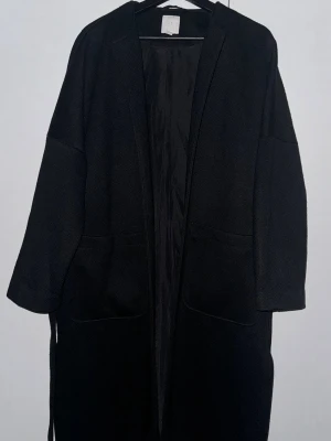 Svart kappa från H&M med bälte - Svart lång kappa från H&M med bälte i midjan och stora fickor framtill. Stilren och enkel design med rak passform och långärmade ärmar. Materialet känns som ullblandning och kappan har en slät insida. Perfekt för dig som gillar minimalistisk stil.