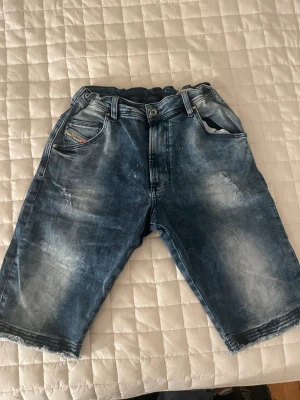 Blå denimshorts med slitningar - Säljer ett par blå denimshorts från Diesel med coola slitningar och tvättade detaljer. Shortsen har klassisk femficksdesign, knapp och dragkedja framtill samt råa kanter vid bensluten. Perfekt för dig som gillar en avslappnad streetstil.