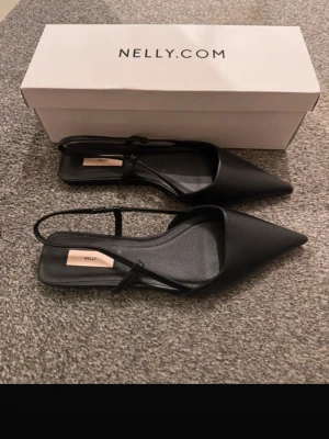 Svarta skor från Nelly - Stilrena svarta ballerinaskor från Nelly med spetsig tå och tunna remmar runt hälen. Skorna har platt sula och är tillverkade i syntetmaterial. Perfekta för dig som gillar minimalistisk och trendig look.