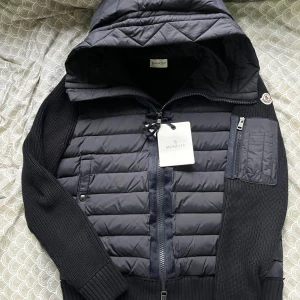 Moncler Cardigan - Snygg Navy Blue Cardigan fràn Moncler med quiltad front och huva, stickade ärmar och rygg. Jackan har dragkedja framtill, ficka med dragkedja pà ärmen och Moncler-logga pả axeln. Perfekt för dig som vill ha en fin och stilren look.