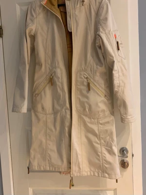 Beige fleecefodrad regnjacka från Ilse Jacobsen - Snygg beige regnjacka från Ilse Jacobsen Cruise med långa ärmar, huva och dragkedja. Jackan har vattentäta tejpade sömmar, har fleece fodrad insida, andasfunktion och praktiska fickor med dragkedja. Perfekt för blöta dagar och har en stilren, lång siluett.